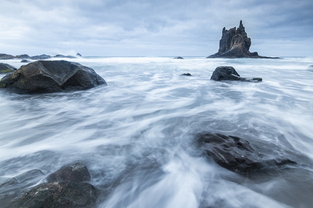 Hermosa foto de agua que fluye alrededor de grandes piedras cerca de la roca de Benijo en un día nublado en España