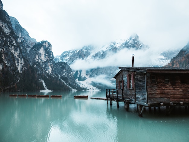 Hermosa foto de una casita de madera junto a un lago en un muelle con increíbles montañas nubladas y nevadas