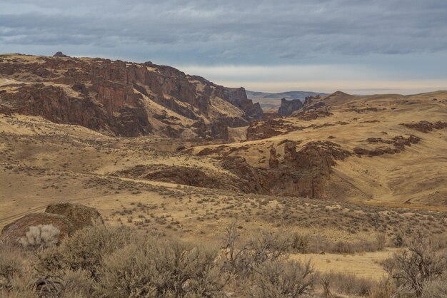 Hermosa foto de un desierto con montañas cubiertas de arbustos secos bajo un cielo nublado azul