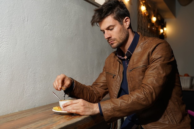 Hermosa foto de un hombre elegante en una chaqueta de cuero marrón agregando azúcar a su café | Foto