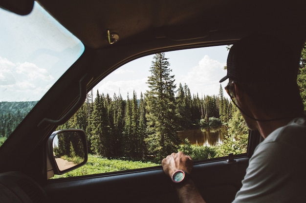 Hermosa foto de un hombre sentado en el coche disfrutando de la vista de los pinos cerca del estanque