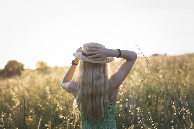 Hermosa foto de una joven rubia con un sombrero de pie en el campo al atardecer