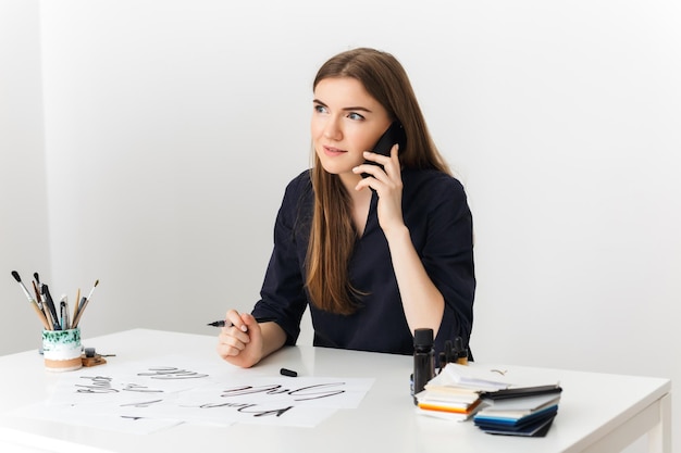 Hermosa foto de una joven sentada en el escritorio blanco y hablando por su celular mientras felizmente mira a un lado aislada
