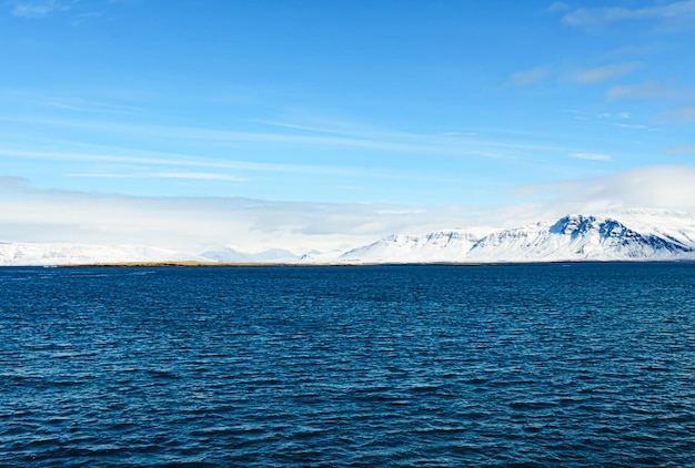 Hermosa foto de un mar azul lleno de olas frente a una montaña nevada en Islandia