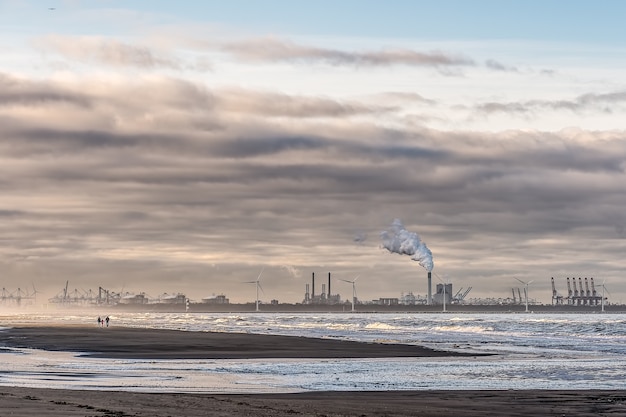 Hermosa foto de un mar con molinos de viento y fábrica en la distancia bajo un cielo nublado