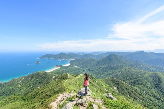 Hermosa foto de una mujer de pie sobre un acantilado con un paisaje de colinas boscosas y un océano azul