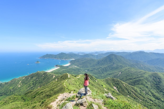 Hermosa foto de una mujer de pie sobre un acantilado con un paisaje de colinas boscosas y un océano azul