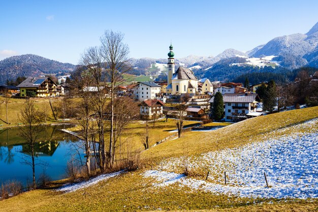 Hermosa foto de un pequeño pueblo rodeado por un lago y colinas nevadas