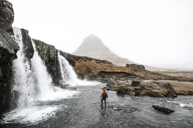 Hermosa foto de una persona de pie en el agua cerca de cascadas que fluyen por las colinas