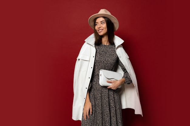 hermosa gir morena europea en chaqueta blanca de moda y vestido con estampado posando. Sosteniendo el bolso de cuero.