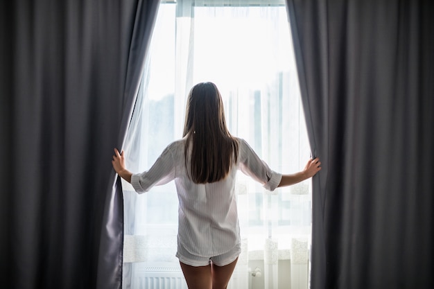 Hermosa joven abriendo cortinas y mirando por la ventana