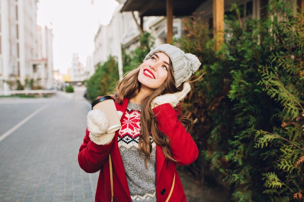 Hermosa joven de abrigo rojo y gorro de punto caminando en la calle. Ella sostiene el café para llevar con guantes blancos, soñando con el cielo.