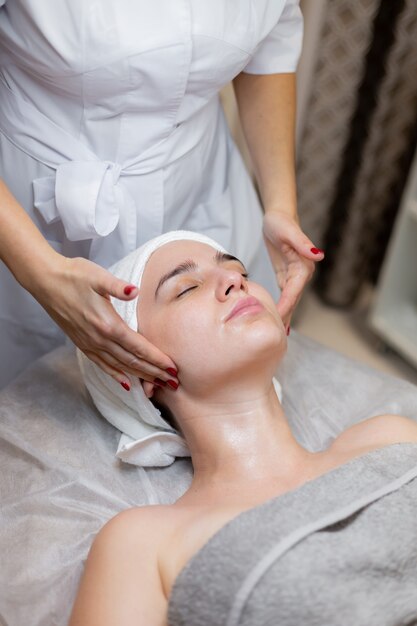 Una hermosa joven se acuesta en la mesa de la esteticista y recibe procedimientos, un ligero masaje facial con aceite.