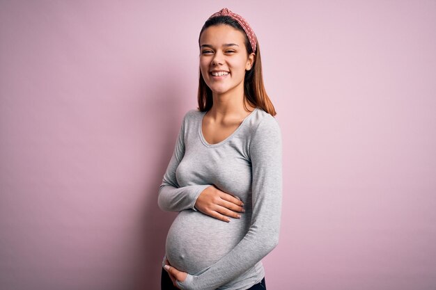 Hermosa joven adolescente embarazada esperando bebé sobre fondo rosa aislado cara feliz sonriendo con los brazos cruzados mirando a la cámara Persona positiva