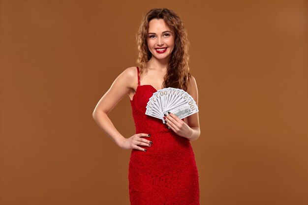 Hermosa joven afortunada feliz mujer de cabello castaño con vestido de cóctel rojo muestra el fanático del dinero de los billetes de 100 dólares, fondo marrón. Éxito, fortuna, lotería.