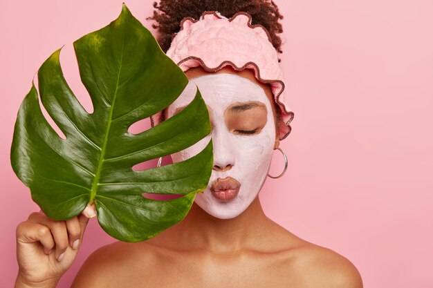 Hermosa joven afro se aplica una máscara de arcilla nutritiva, tiene los ojos cerrados, los labios doblados, cubre la mitad de la cara con una gran hoja verde, usa una diadema de ducha, aislada en una pared rosa