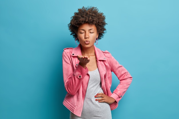 Hermosa joven afroamericana romántica envía apasionado beso al aire, mantiene las palmas hacia adelante, los labios doblados, sopla mwah, vestida con ropa de moda, cierra los ojos