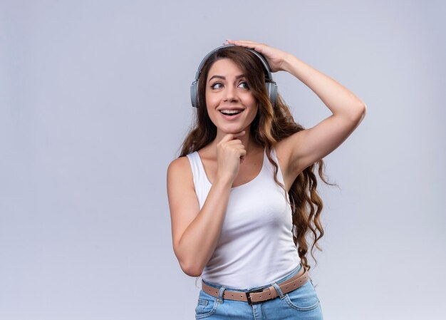 Hermosa joven alegre con audífonos poniendo las manos en la cabeza y debajo de la barbilla mirando hacia el lado derecho con espacio de copia