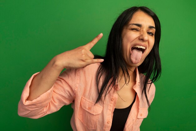 Hermosa joven alegre con camiseta rosa mostrando la lengua haciendo gesto de cabra aislado en la pared verde