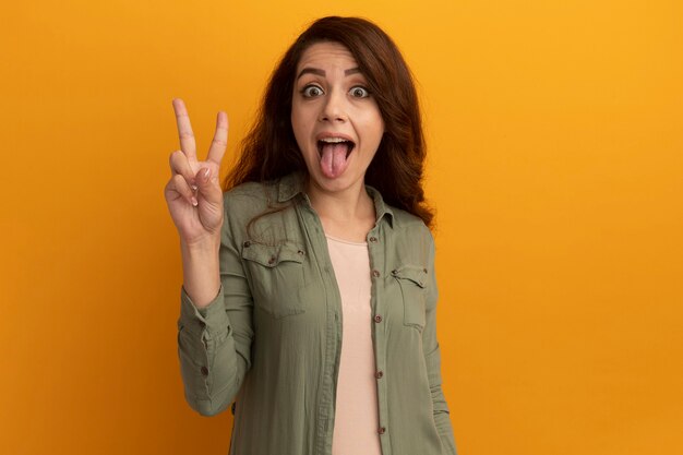 Hermosa joven alegre con camiseta verde oliva mostrando la lengua y haciendo gesto de paz aislado en la pared amarilla con espacio de copia