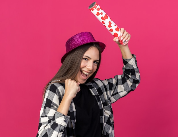 Hermosa joven alegre con gorro de fiesta sosteniendo cañón de confeti mostrando gesto de sí