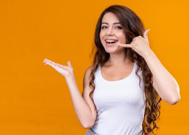 Hermosa joven alegre haciendo distintivo de llamada y mostrando la mano vacía en el espacio naranja aislado