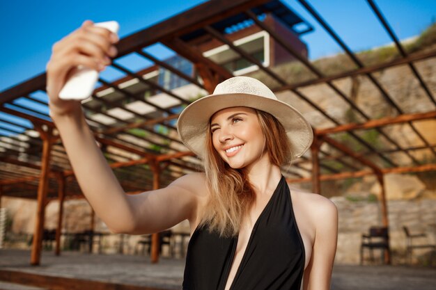 Hermosa joven alegre con sombrero descansa en la playa de la mañana y hace selfie