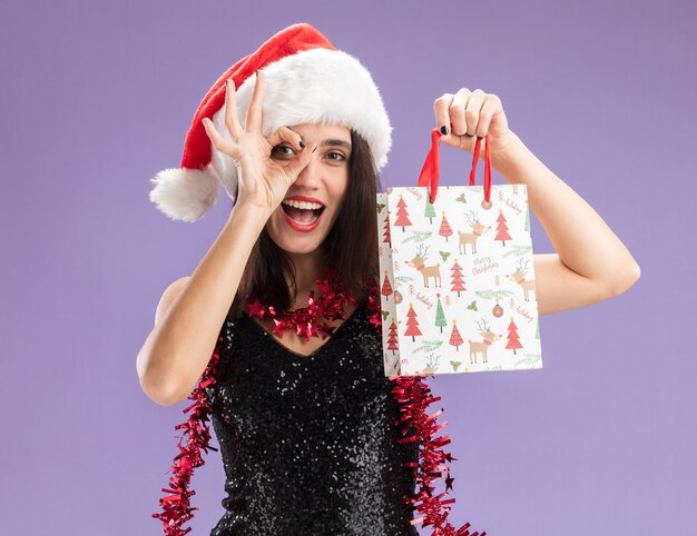 Hermosa joven alegre con sombrero de navidad con guirnalda en el cuello sosteniendo una bolsa de regalo mostrando gesto de mirada aislado sobre fondo púrpura