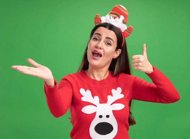 Hermosa joven alegre vistiendo un suéter de navidad con puntos de aro de pelo de navidad con la mano al lado aislado en la pared verde