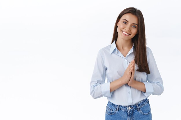 Hermosa joven de apariencia amigable lista para ayudar al cliente con cualquier problema, juntar las manos cerca del pecho e inclinar la cabeza escuchando con placer, sonriendo, diciendo por favor o agradeciendo el favor.