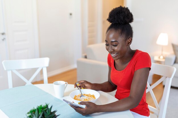 Hermosa joven arfican americana enviando mensajes de texto en un teléfono inteligente en casa Señora alegre comiendo copos de maíz bebiendo café por la mañana y mirando su teléfono móvil