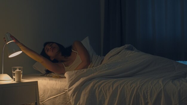 Hermosa joven asiática durmiendo cómodamente en su habitación. Mano femenina india que apaga el interruptor de la luz cerca de la cama en la habitación en casa a altas horas de la noche listo para dormir. Dulces sueños y relajación.