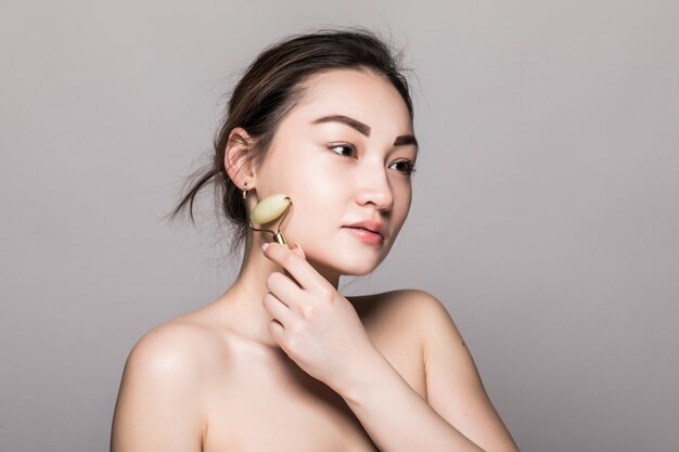 Hermosa joven asiática con un rodillo de cara de jade en su piel perfecta. Primer plano de cara de belleza. Conceptual de tratamientos faciales con piedras semipreciosas. aislado en gris con espacio de copia