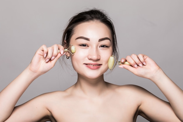 Hermosa joven asiática con un rodillo de cara de jade en su piel perfecta. Primer plano de cara de belleza. Conceptual de tratamientos faciales con piedras semipreciosas. aislado en gris con espacio de copia