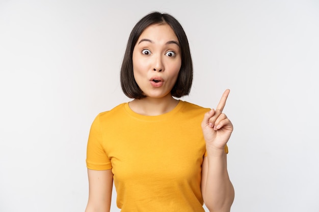 Hermosa joven asiática señalando con el dedo sonriendo y mirando divertida a la cámara que muestra un anuncio publicitario en el fondo blanco superior