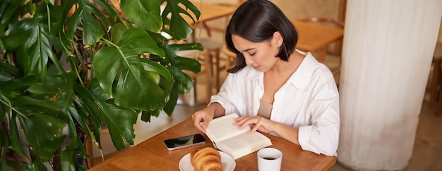 Hermosa joven asiática sentada en un café con un libro comiendo croissant y leyendo bebiendo una taza de