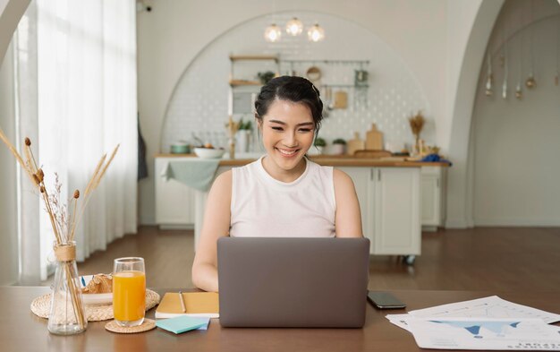 Hermosa joven asiática trabajando en una computadora portátil mientras se sienta en el fondo de la sala de la cocina