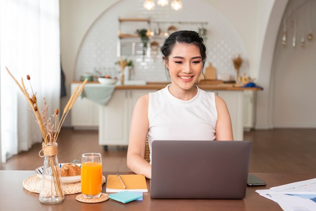 Hermosa joven asiática trabajando en una computadora portátil mientras se sienta en el fondo de la sala de la cocina