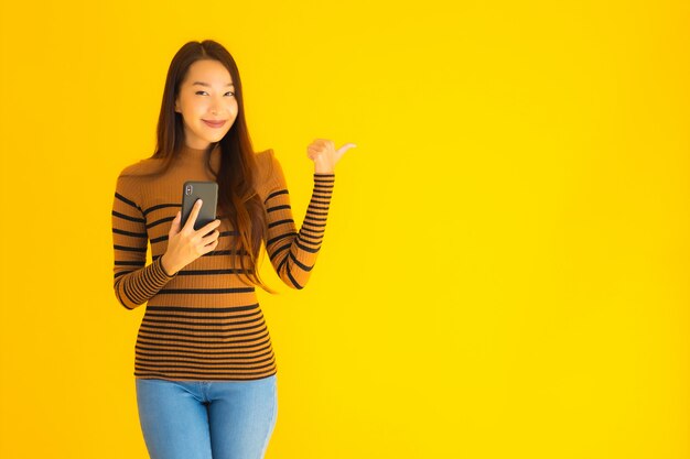 Hermosa joven asiática utiliza teléfono móvil inteligente o teléfono celular con mucha acción en la pared amarilla