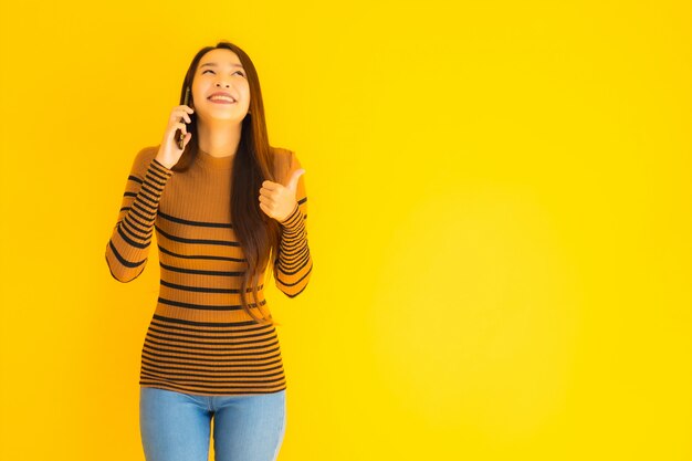 Hermosa joven asiática utiliza teléfono móvil inteligente o teléfono celular con mucha acción en la pared amarilla