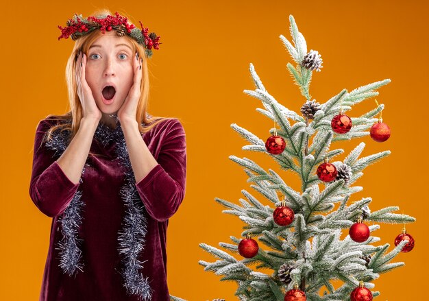 Hermosa joven asustada de pie cerca del árbol de Navidad con vestido rojo y corona con guirnalda en el cuello poniendo las manos en las mejillas aisladas sobre fondo naranja