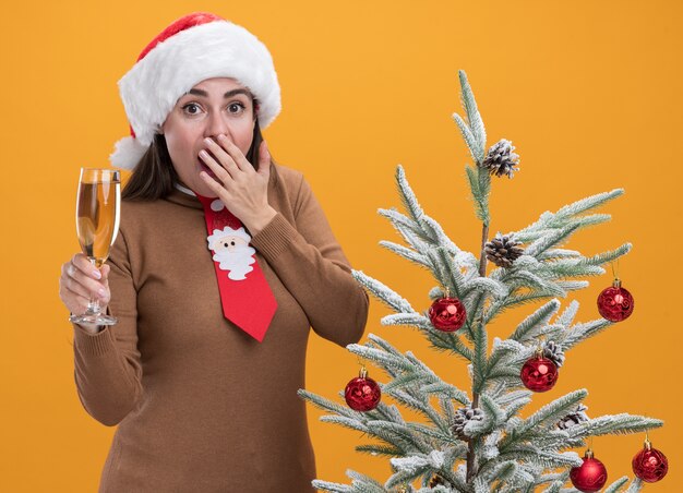 Hermosa joven asustada con sombrero de Navidad con corbata de pie cerca del árbol de Navidad sosteniendo una copa de champán con la boca cubierta con la mano aislada en la pared naranja