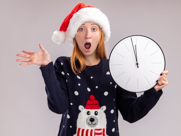 Hermosa joven asustada con sombrero de navidad sosteniendo el reloj de pared extendiendo la mano aislada en la pared blanca