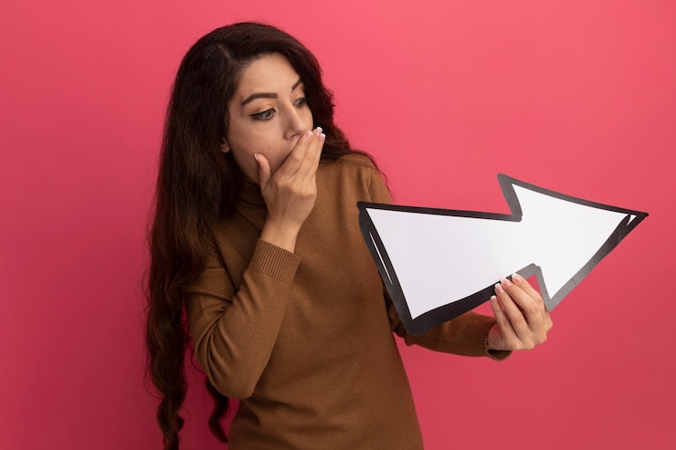 Foto gratuito hermosa joven asustada con suéter de cuello alto sosteniendo y mirando la marca de dirección aislada en la pared rosa