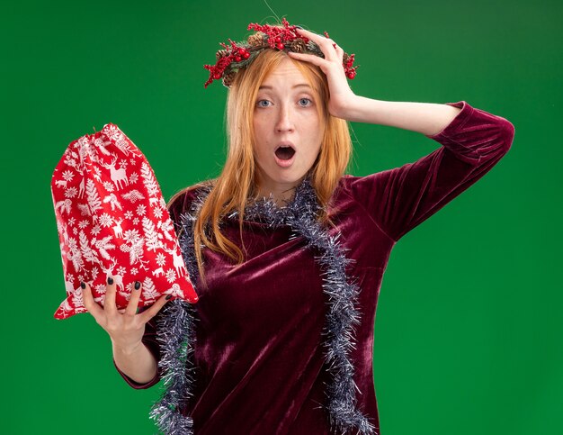 Hermosa joven asustada con vestido rojo con corona y guirnalda en el cuello sosteniendo una bolsa de Navidad poniendo la mano en la cabeza aislada sobre fondo verde