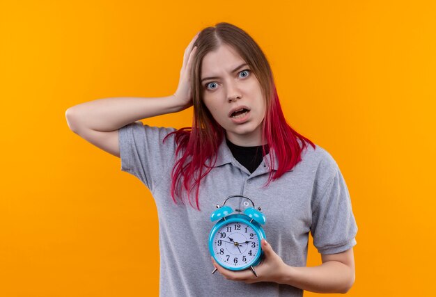 Hermosa joven asustada vistiendo camiseta gris sosteniendo el despertador poniendo la mano en la cabeza sobre fondo amarillo aislado