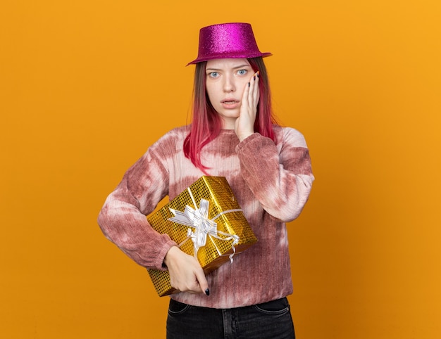 Hermosa joven asustada vistiendo gorro de fiesta con caja de regalo poniendo la mano en la mejilla