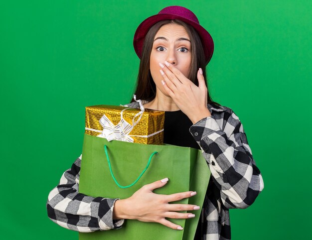 Hermosa joven asustada vistiendo gorro de fiesta sosteniendo una bolsa de regalo con la boca cubierta con la mano