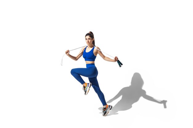 Hermosa joven atleta practicando sobre fondo blanco de estudio, retrato con sombras. Modelo de ajuste deportivo en movimiento y acción.