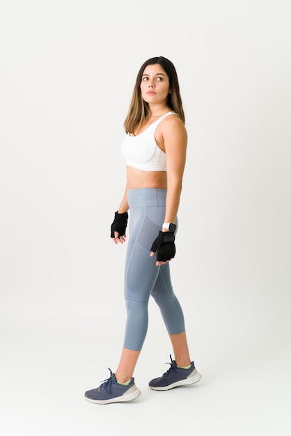 Hermosa joven atleta en ropa activa usando guantes para levantar pesas en el gimnasio. Deportiva mujer caucásica contra un fondo blanco.